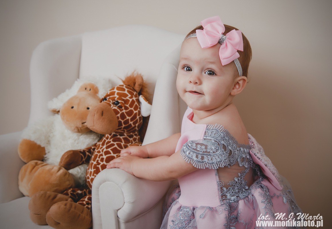 Uśmiechnięta dziewczynka w różowej sukience z koronkowymi aplikacjami i kokardą na głowie, siedząca w białym fotelu obok pluszowej owieczki i żyrafy.