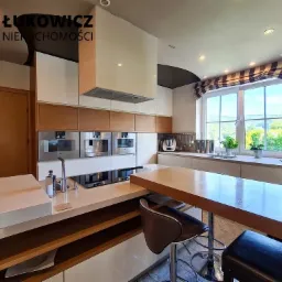 Nowoczesna kuchnia z drewnianymi akcentami, wyspą z hokerami i oknem z widokiem na zieleń, w tle wbudowane urządzenia AGD.