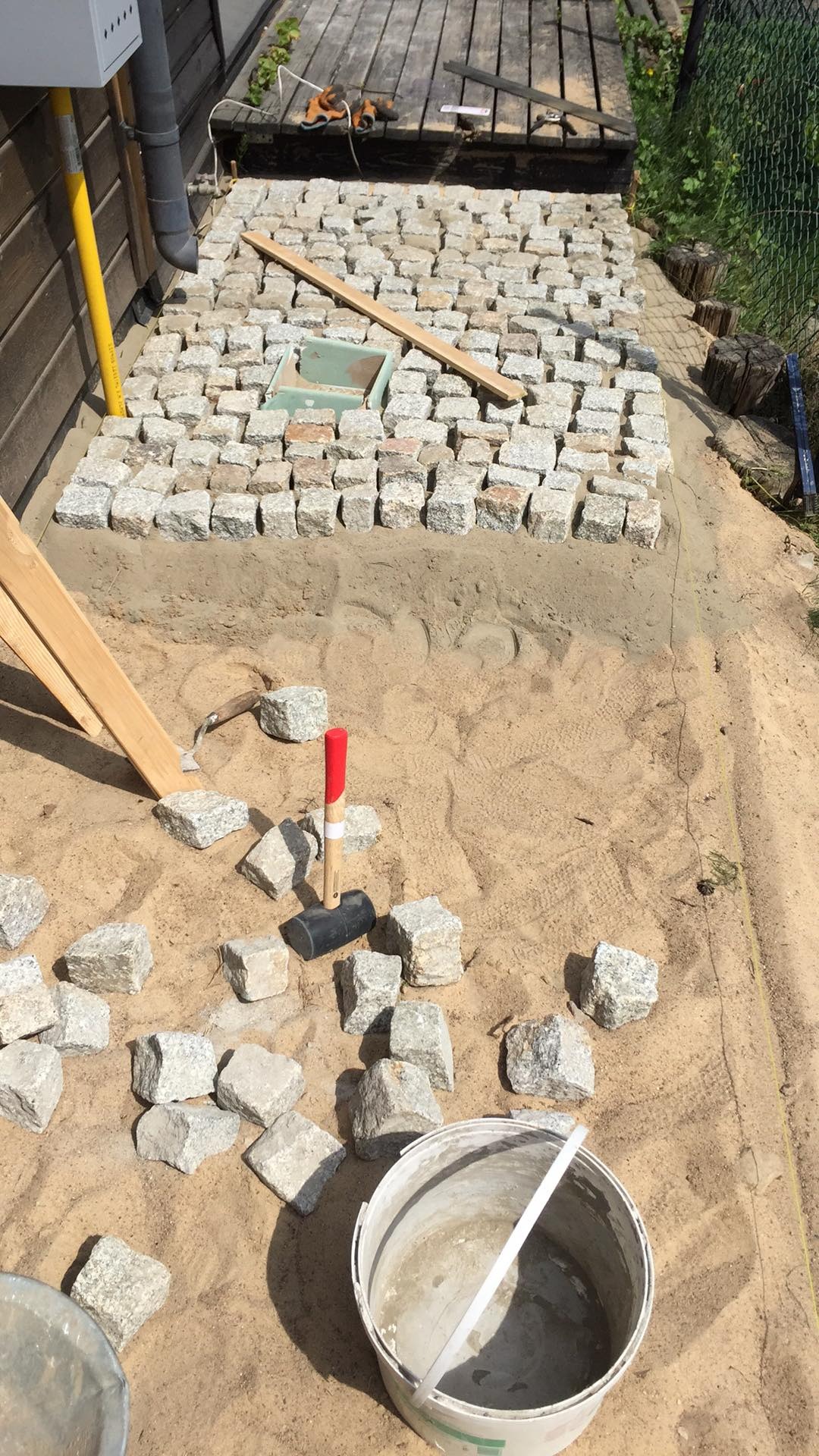 Układanie kostki granitowej na podsypce piaskowej, widoczny fragment ułożonej nawierzchni, narzędzia pracy: młotek brukarski, poziomica, sznur traserski oraz wiadro z zaprawą.