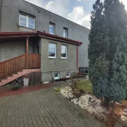 Szary, dwukondygnacyjny dom z drewnianym gankiem i schodami, widok na elewację z oknami i czerwonymi rynnami, obok rosną duże drzewa iglaste.