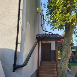 Świeżo otynkowana elewacja budynku z brązowym zadaszeniem nad wejściem, widoczne rynny i fragment drzewa.