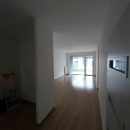 Wnętrze pustego, jasnego mieszkania po remoncie, z drewnianą podłogą i białymi ścianami, widok na taras przez przeszklone drzwi balkonowe, drabina malarska w rogu.