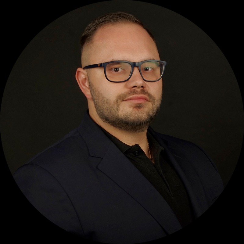 Portret mężczyzny w ciemnym garniturze i okularach na czarnym tle, z delikatnym zarostem i widocznym złotym łańcuszkiem pod koszulą.