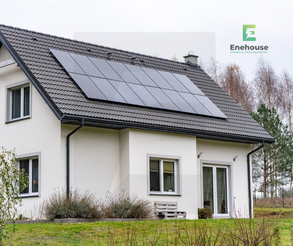 Biały dom jednorodzinny z szarym dachem, na którym zamontowano panele fotowoltaiczne. Widoczne okna, drzwi balkonowe i ogród.