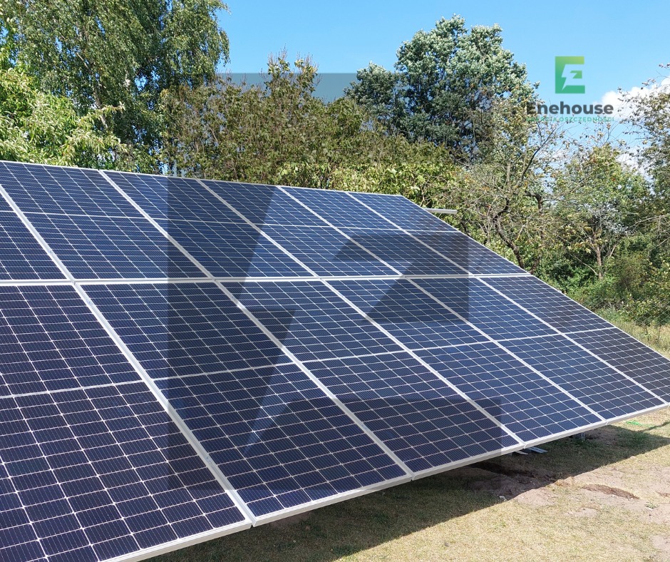 Instalacja fotowoltaiczna na trawiastym terenie, widoczna pod kątem. Panele w ciemnych odcieniach z białymi ramami. W tle drzewa i błękitne niebo.