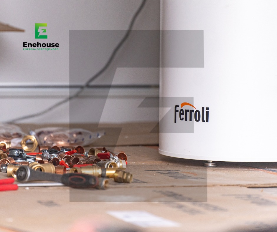 Biały zasobnik ciepłej wody Ferroli z armaturą miedzianą i narzędziami na kartonie, widoczne logo firmy Enehouse w tle. Instalacja grzewcza w trakcie montażu.