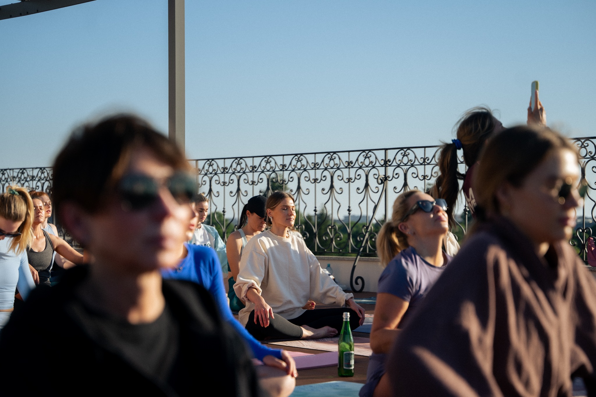 Grupa osób ćwiczących jogę na dachu budynku w słoneczny dzień, widoczne maty do ćwiczeń i butelka wody mineralnej, w tle barierka i fragment miasta.