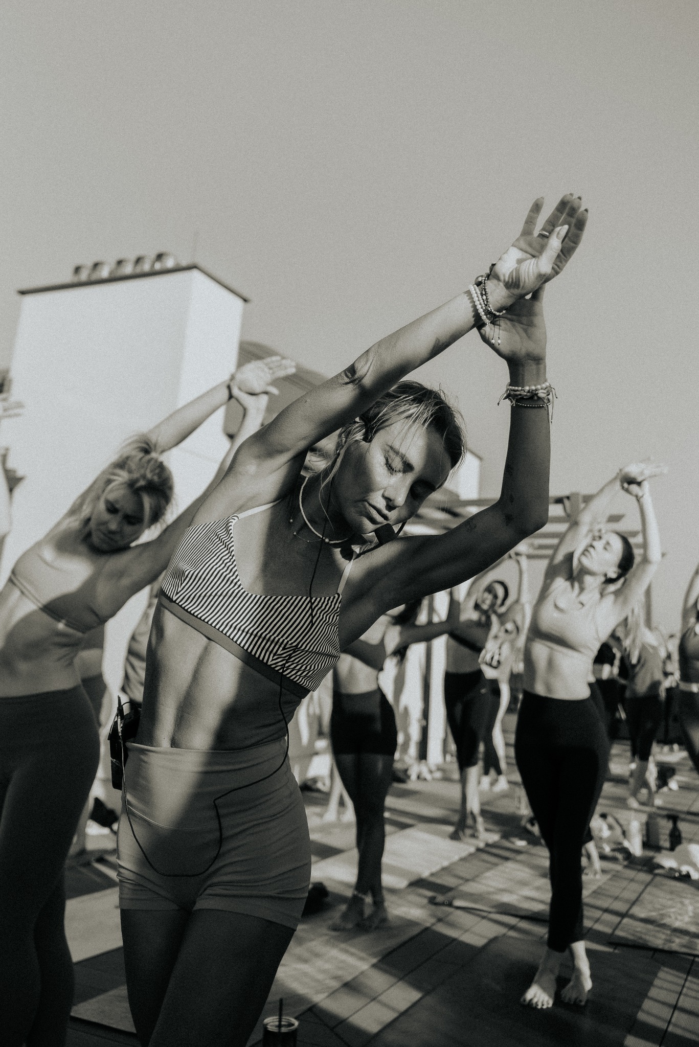 Czarno-białe zdjęcie grupy kobiet wykonujących rozciąganie boczne na dachu budynku w słoneczny dzień. Kobieta na pierwszym planie ma zamknięte oczy, biustonosz w paski i krótkie spodenki.
