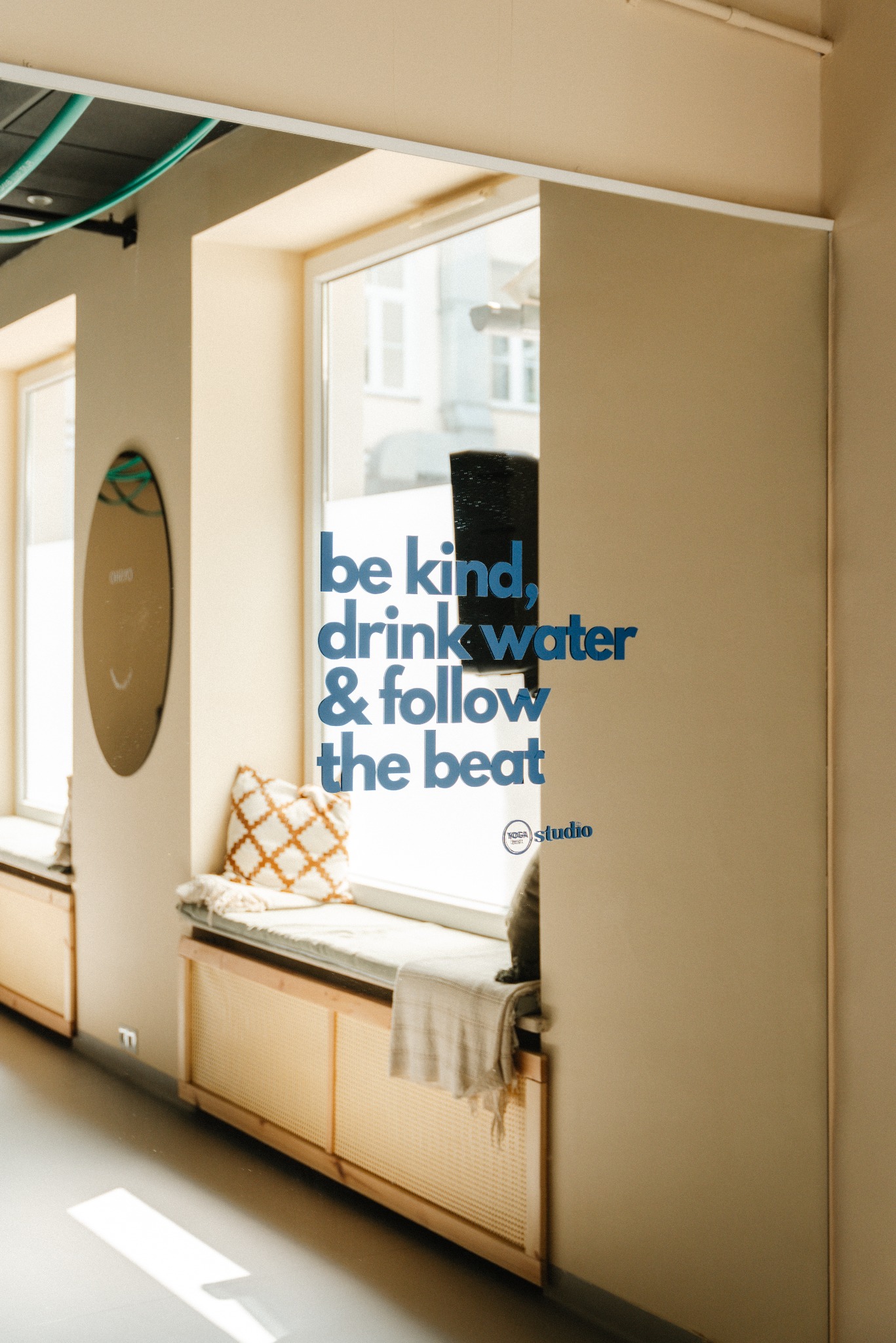 Jasne wnętrze studia z siedziskiem pod oknem, na szybie naklejka z napisem 'be kind, drink water & follow the beat', obok owalne lustro.