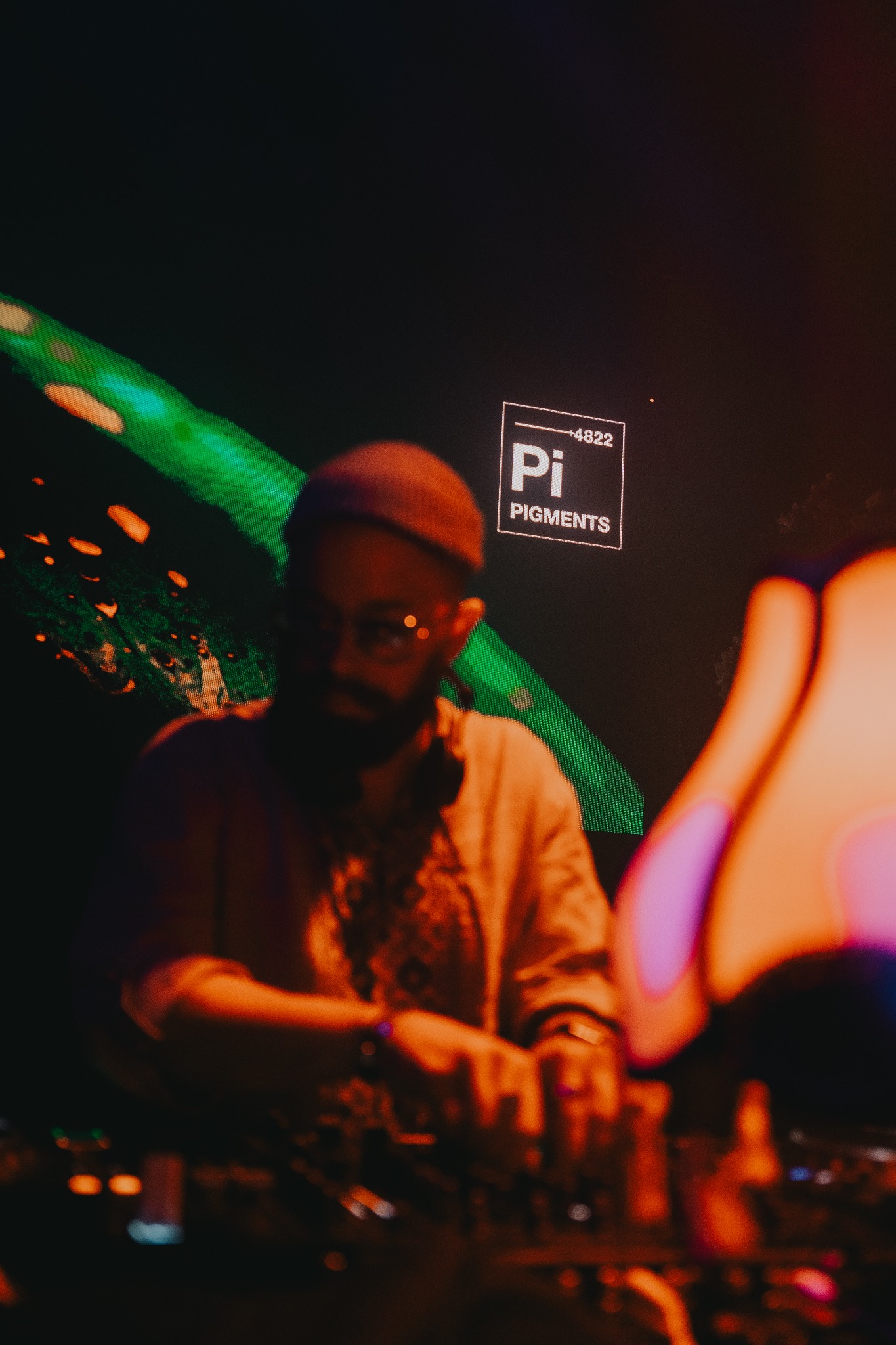 DJ z brodą i okularami w czapce, miksuje muzykę przy konsolecie, w tle neonowe światła i logo 'Pi Pigments'.