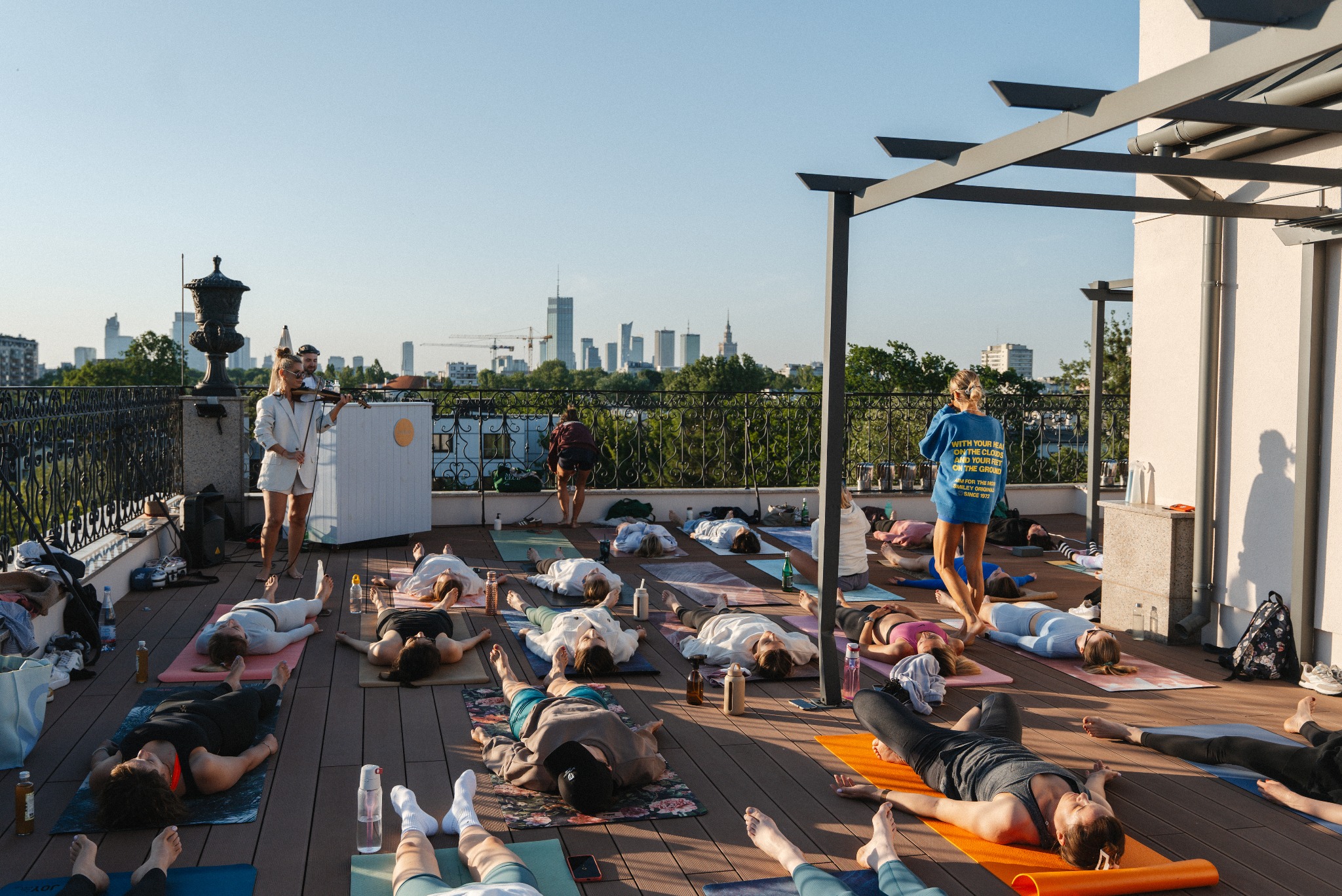 Grupa osób relaksuje się na matach do jogi na tarasie z widokiem na panoramę miasta, przy akompaniamencie skrzypiec