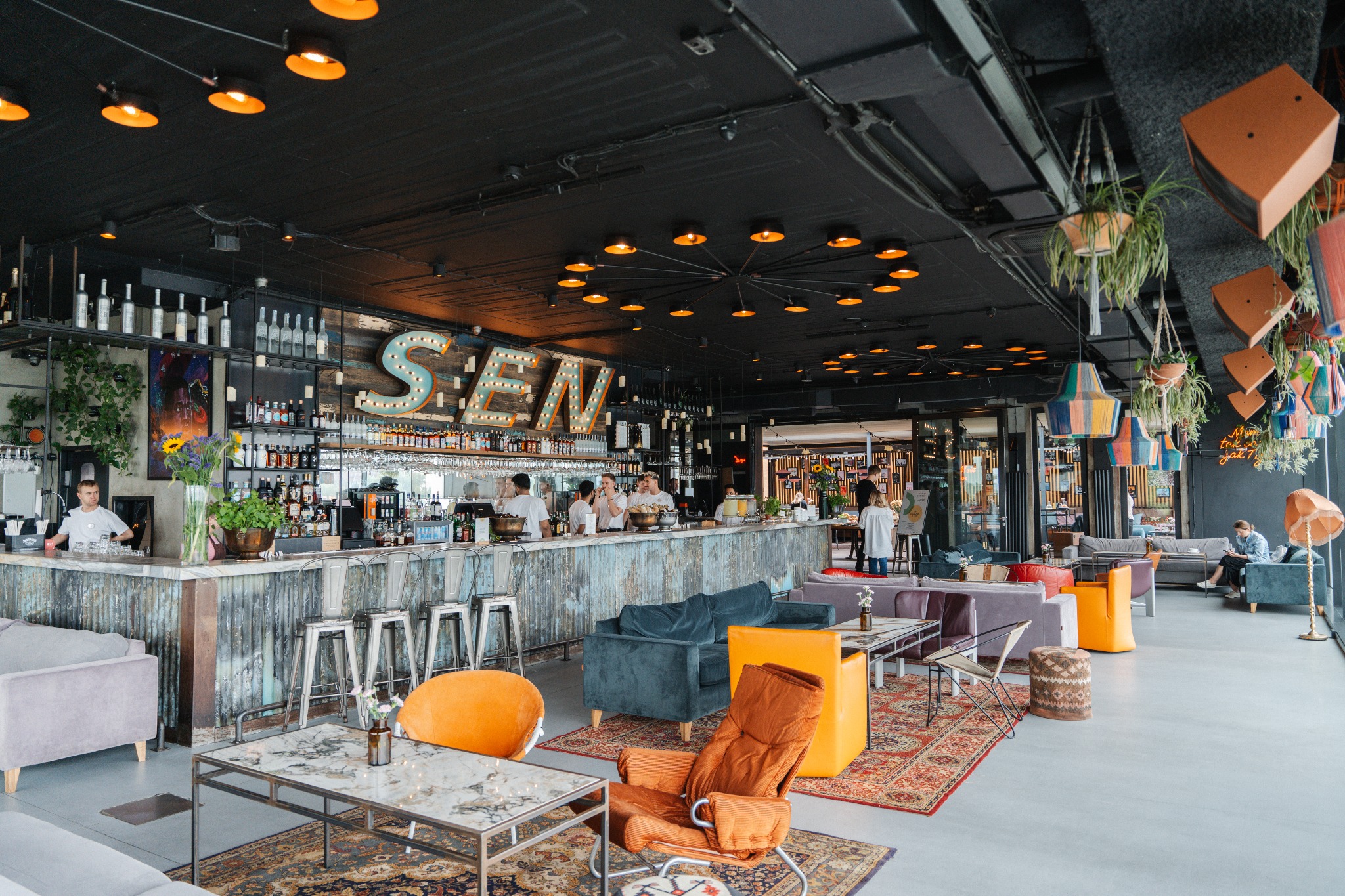 Wnętrze baru z industrialnym wystrojem: blaszany bar podświetlony żarówkami, metalowe stołki barowe, kolorowe sofy i fotele, dywany orientalne, rośliny wiszące w doniczkach, czarny sufit z lampami...