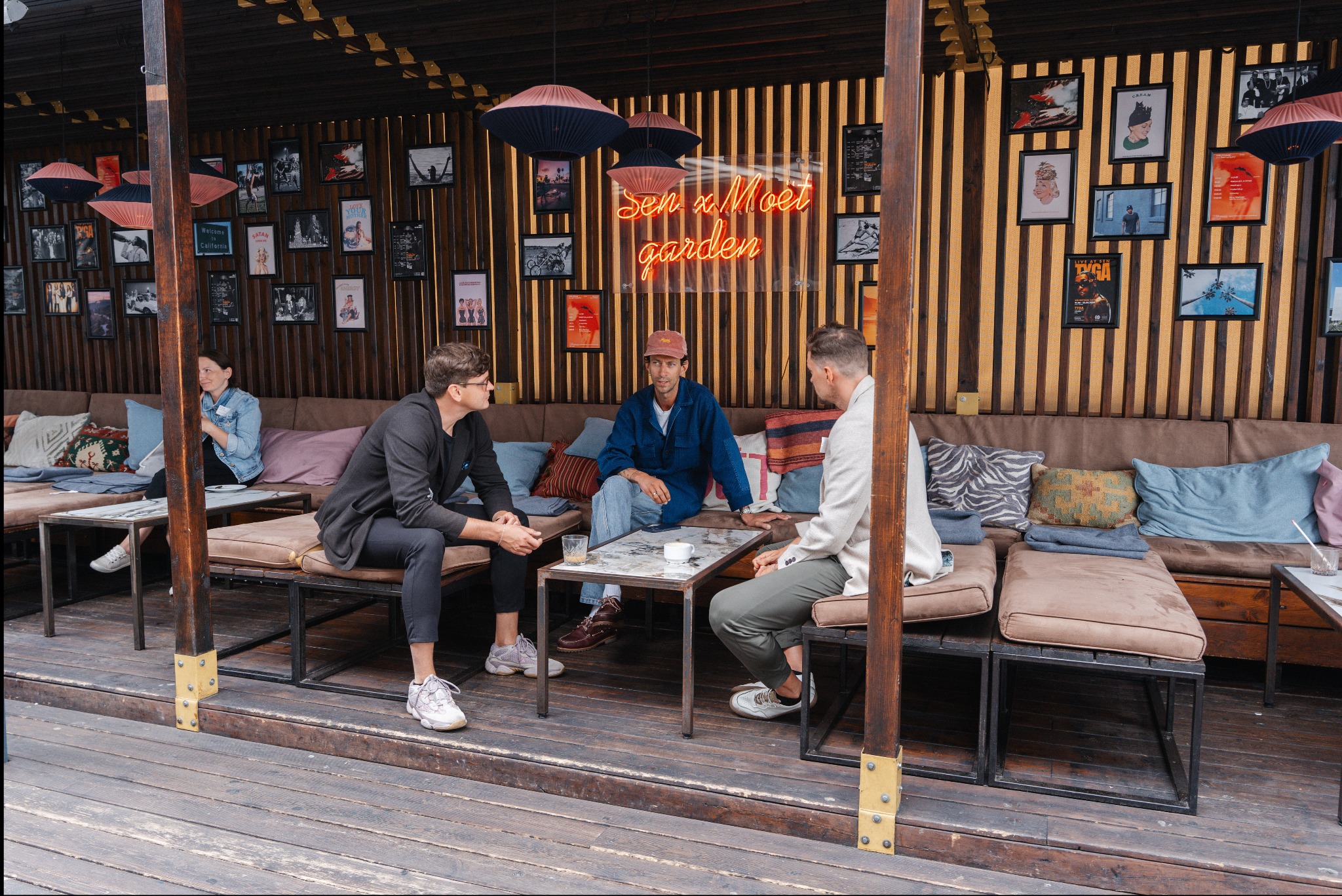 Trzech mężczyzn siedzi przy stoliku na zewnątrz kawiarni z neonowym napisem 'Sen x Moet garden' w tle, otoczeni dekoracyjnymi lampami i ramkami ze zdjęciami na drewnianej ścianie.
