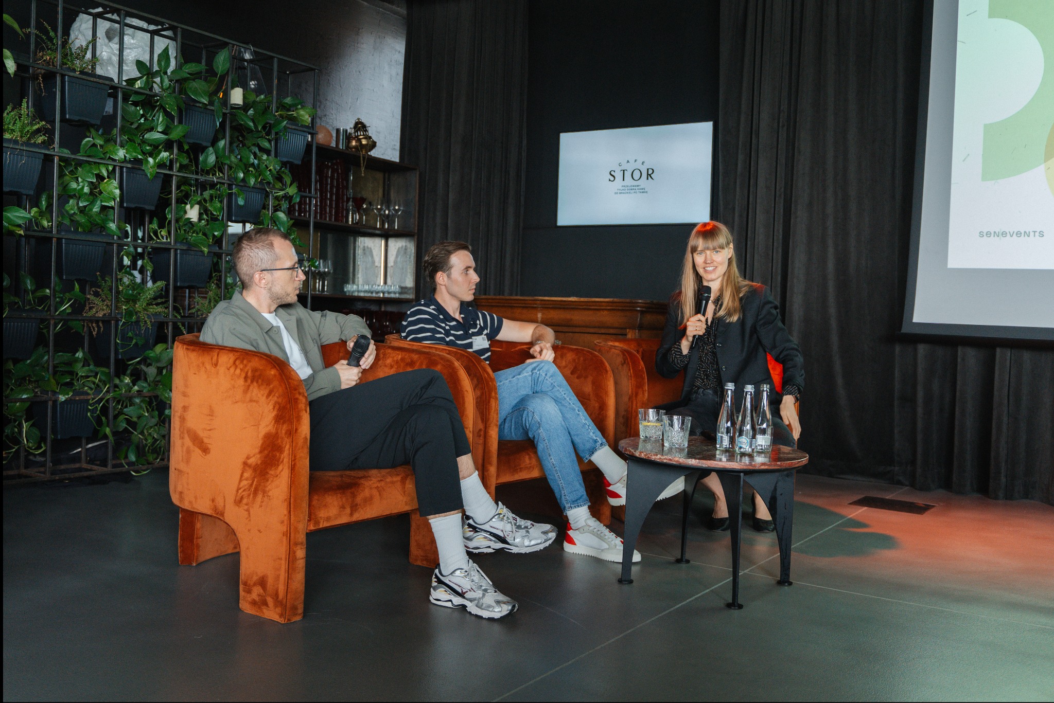 Panel dyskusyjny z trzema osobami siedzącymi w pomarańczowych fotelach na tle ściany z roślinami i ekranu projekcyjnego z logo Cafe Stor.
