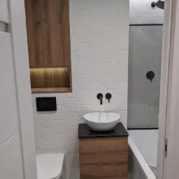 Nowoczesna łazienka z umywalką nablatową, drewnianą szafką pod umywalką, wiszącą szafką z oświetleniem, toaletą i kabiną prysznicową z czarną armaturą na tle białych heksagonalnych płytek.