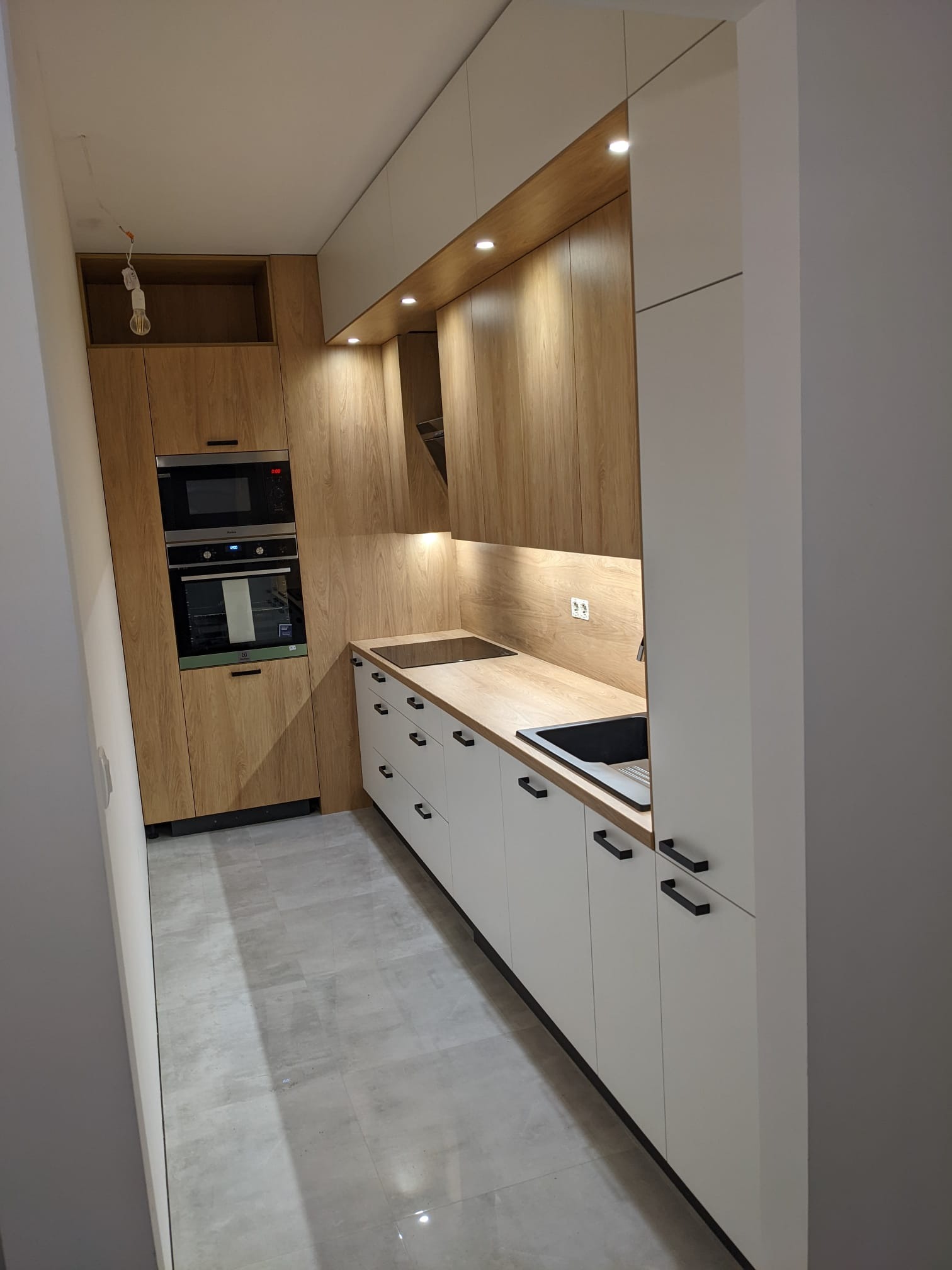 Wąska kuchnia z zabudową w odcieniach bieli i drewna, z czarnymi uchwytami szafek, oświetleniem podszafkowym i sprzętem AGD w zabudowie, widok z perspektywy wejścia do pomieszczenia.