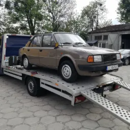 Brązowy samochód Skoda na srebrnej platformie lawety z rozłożonymi najazdami na betonowym placu, widoczny budynek z szarymi bramami w tle.