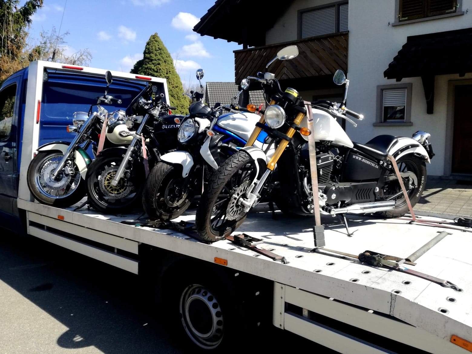 Kilka motocykli różnych marek zabezpieczonych pasami transportowymi na platformie lawety, zaparkowanej przed budynkiem mieszkalnym w słoneczny dzień.