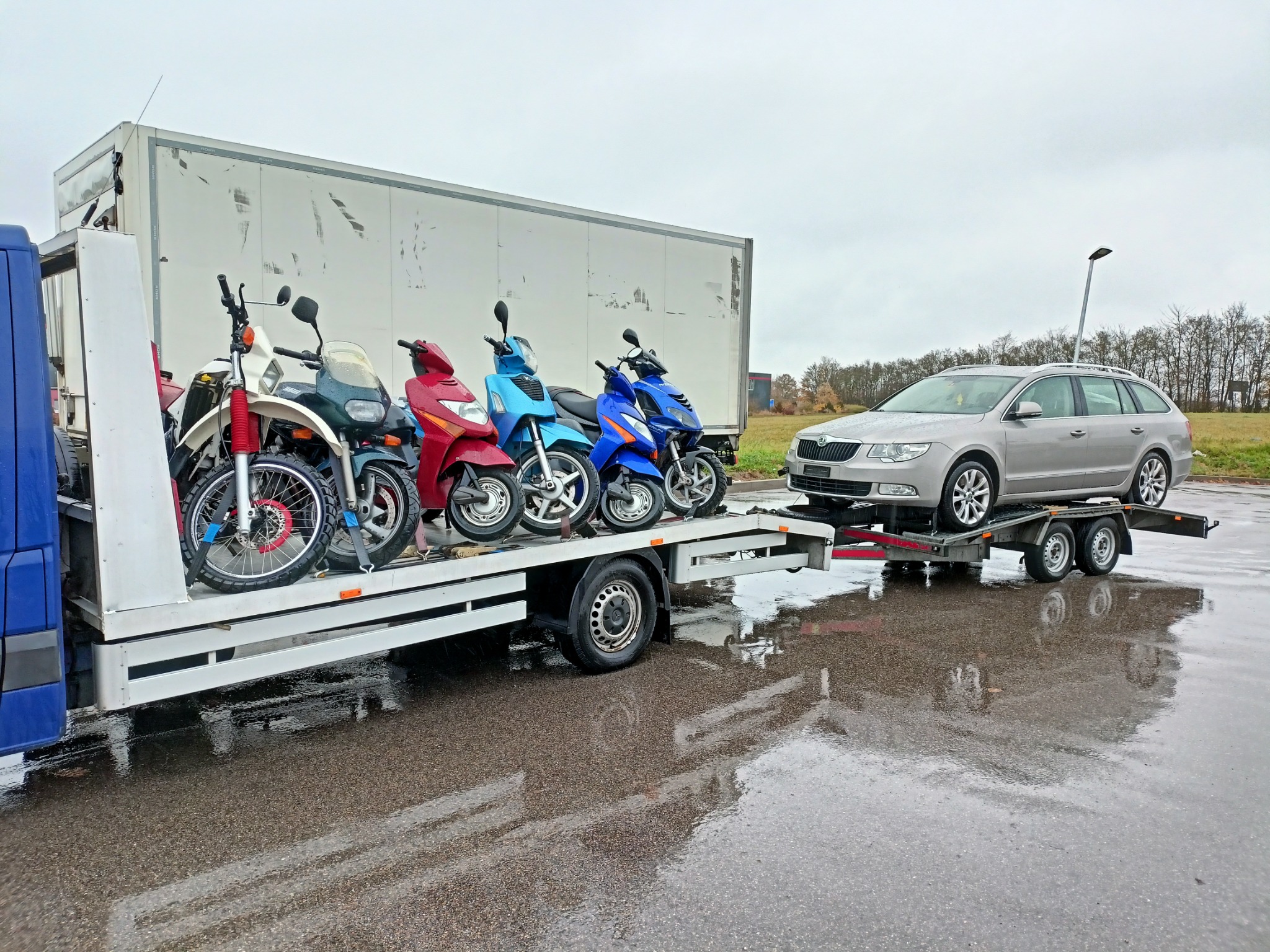 Laweta z załadowanymi motocyklami i samochodem osobowym Skoda na przyczepie, widok z boku na mokrej nawierzchni w pochmurny dzień.