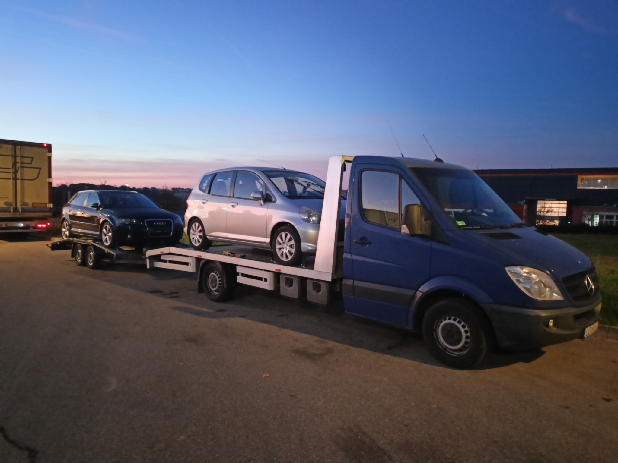 Niebieska laweta Mercedes-Benz z dwoma samochodami (Audi A3 i Honda Jazz) na platformie, sfotografowana o zmierzchu.