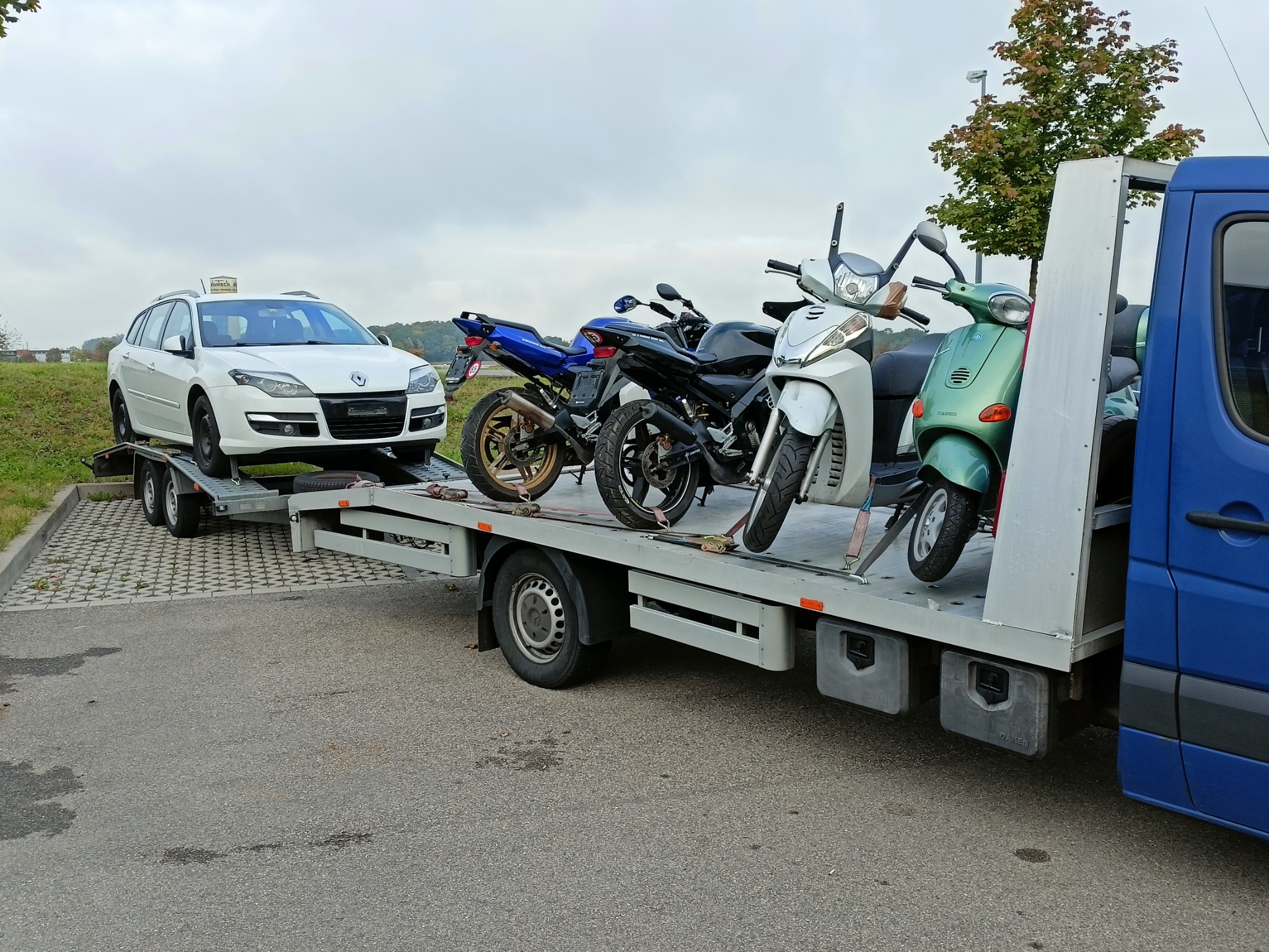 Niebieska laweta z platformą przewożąca białe kombi Renault Laguna oraz trzy motocykle i skuter: niebieski, czarny i zielony Piaggio Vespa.