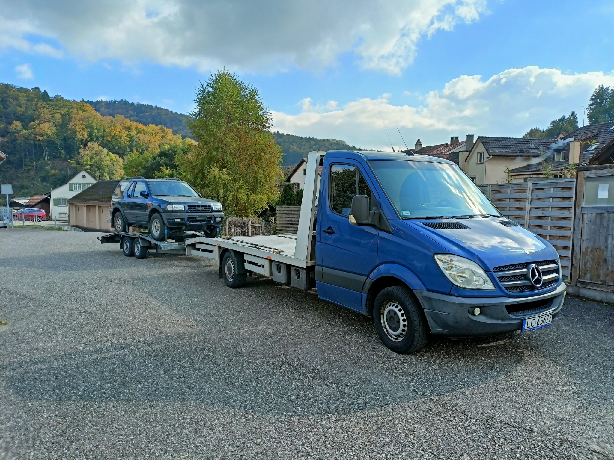Niebieski samochód dostawczy Mercedes-Benz z platformą i przyczepą przewożący ciemny samochód osobowy Opel Frontera na tle zabudowań i drzew.