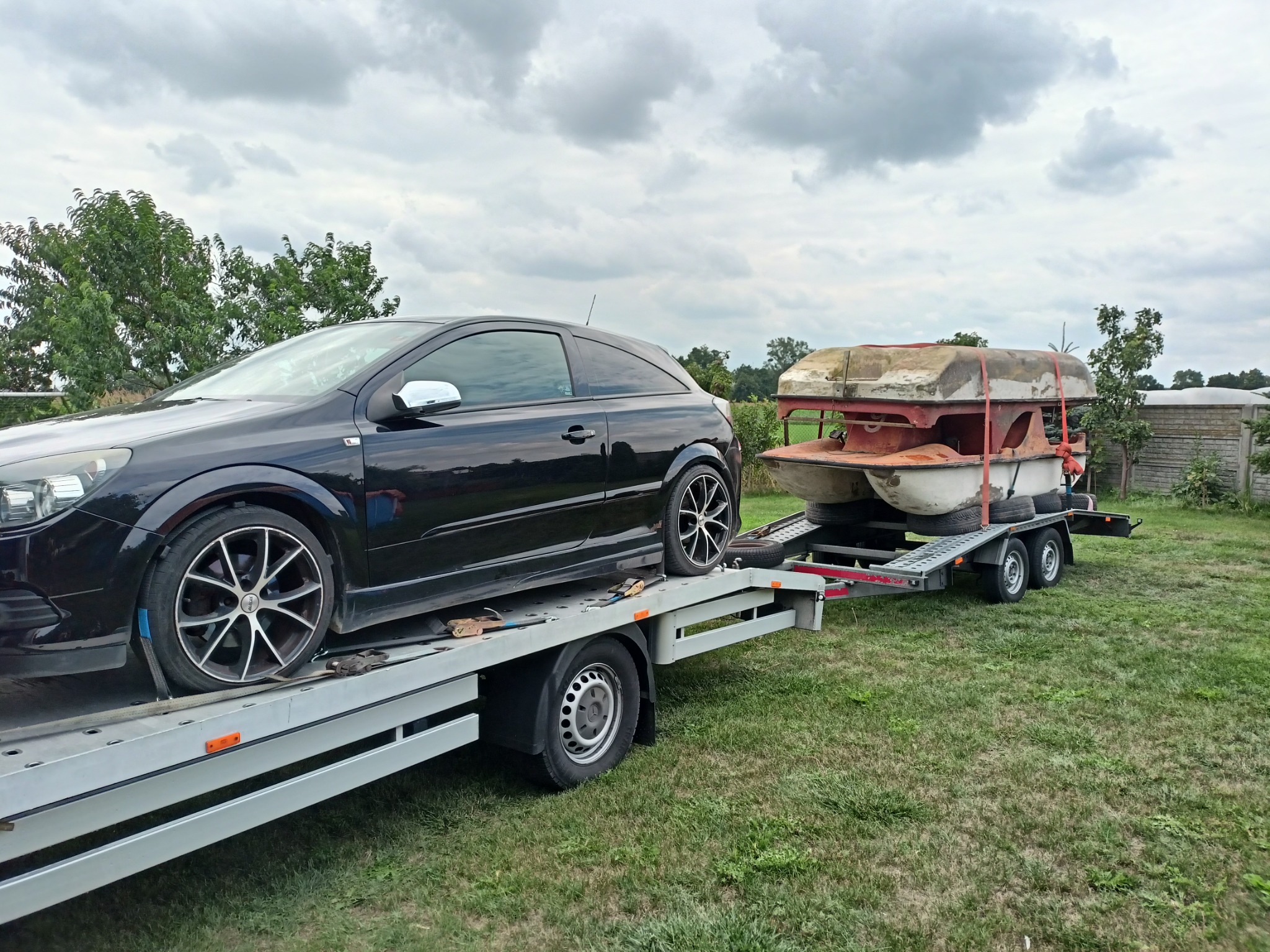 Czarny Opel Astra na lawecie wraz z przyczepą z łódką, transportowany na tle zielonego trawnika i pochmurnego nieba.