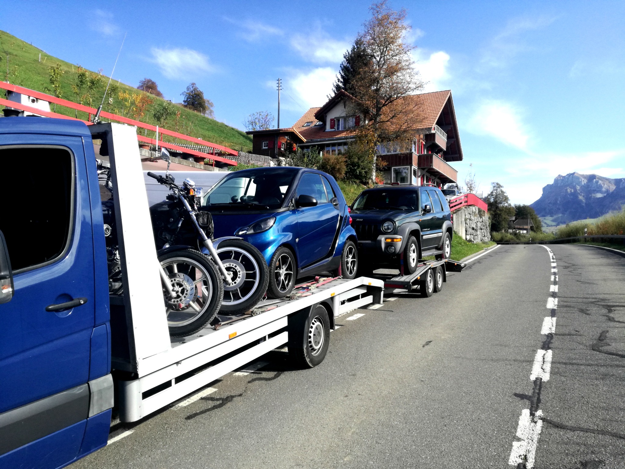 Laweta transportuje motocykl, niebieskiego Smarta i ciemnego Jeepa na tle malowniczego krajobrazu z domem i górą.