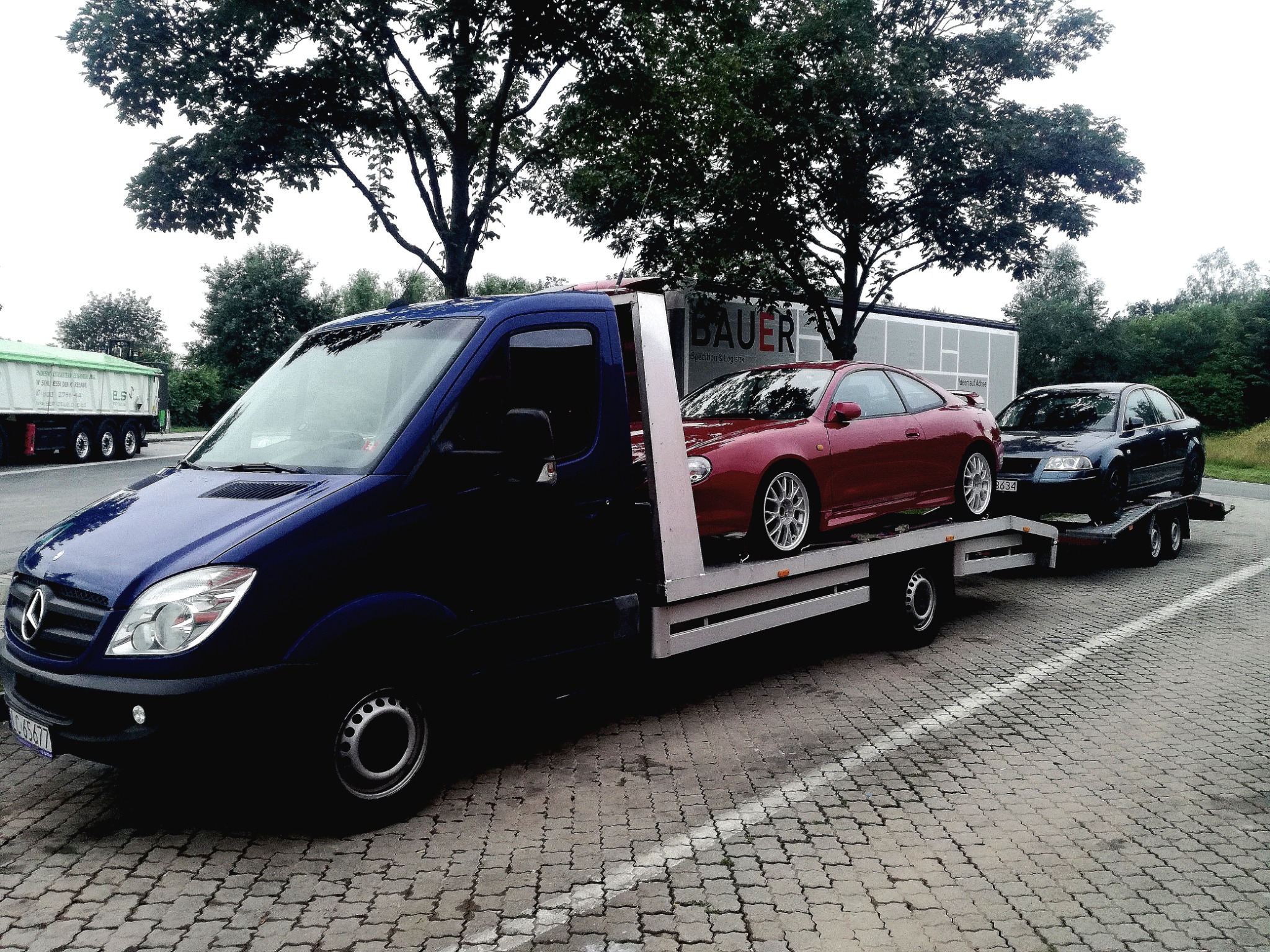 Niebieska laweta Mercedes-Benz z czerwonym sportowym coupe na platformie oraz czarnym sedanem na przyczepie, zaparkowana na brukowanym placu, w tle budynek z napisem BAUER Spedition & Logistik.