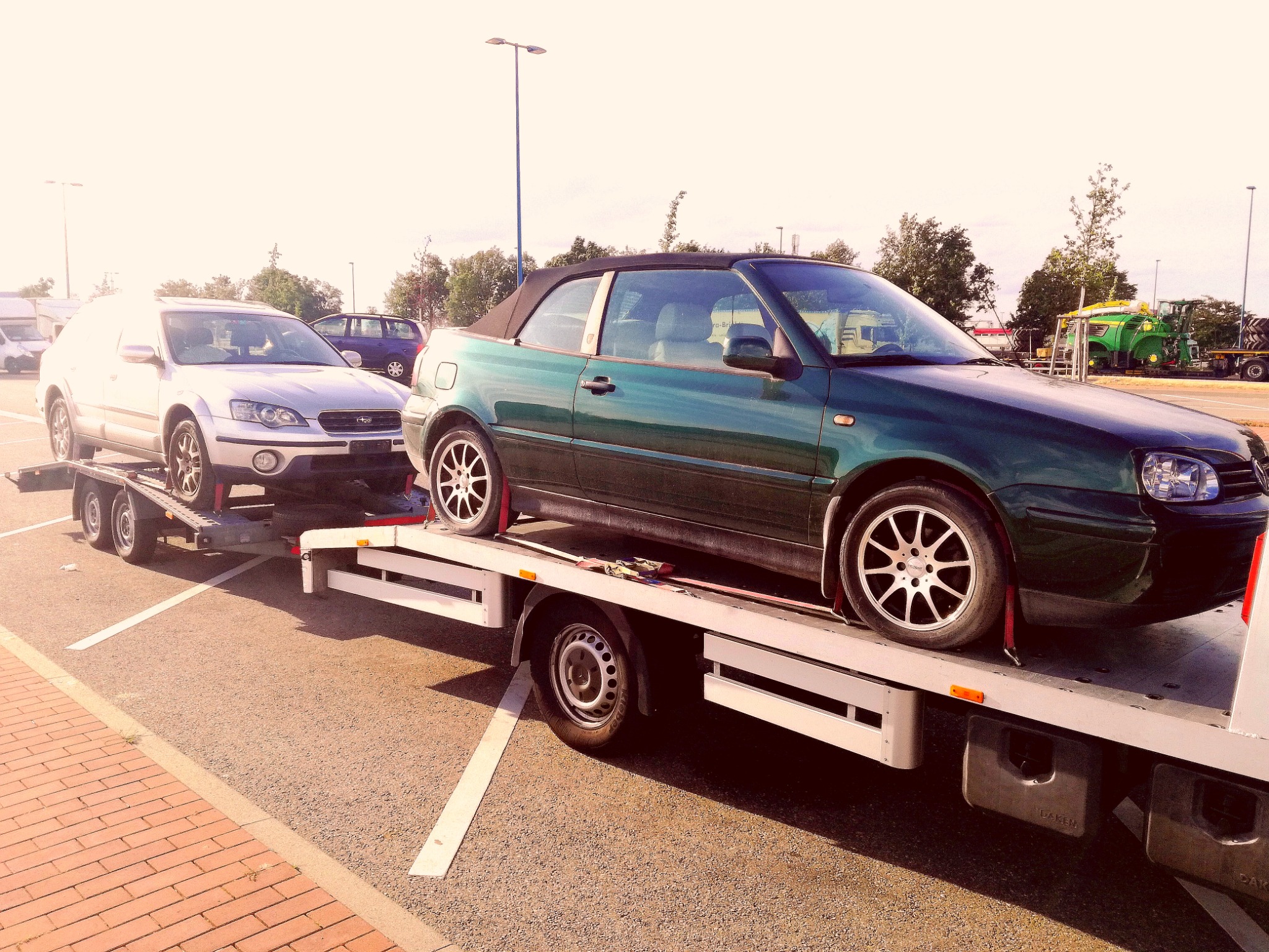 Zielony kabriolet i srebrny samochód osobowy na białej lawecie na parkingu, widoczne zabezpieczenia transportowe na kołach.