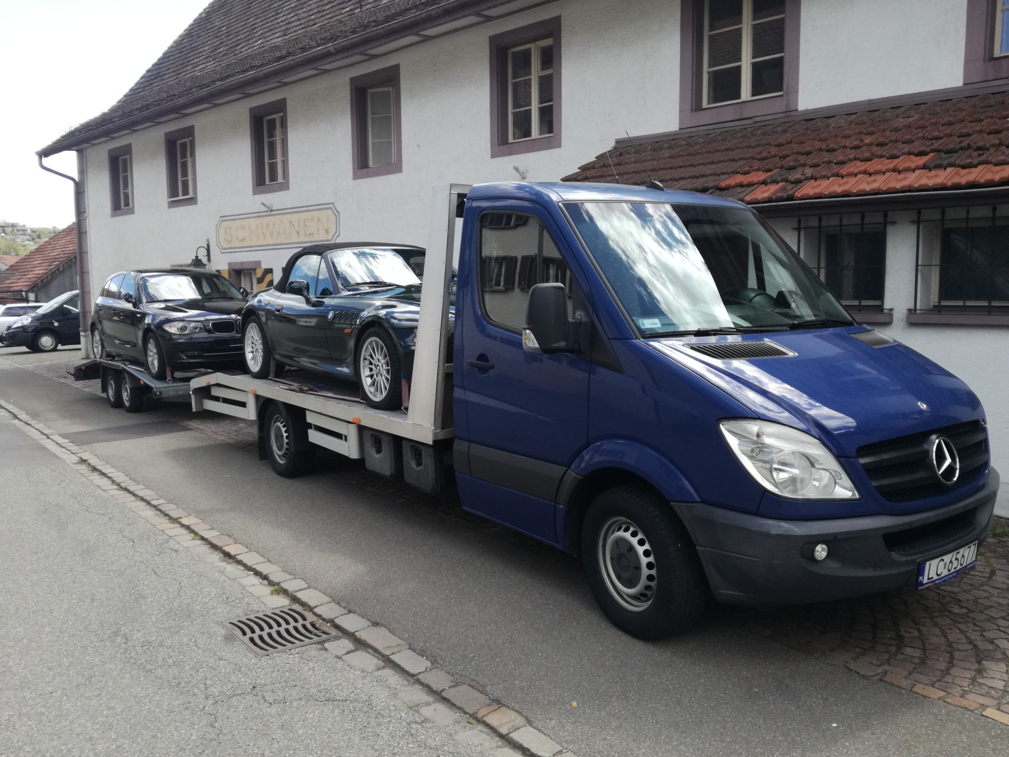 Niebieska laweta Mercedes-Benz Sprinter z dwoma przewożonymi czarnymi samochodami BMW na przyczepie w tle budynku z szyldem 'Schwanen'.