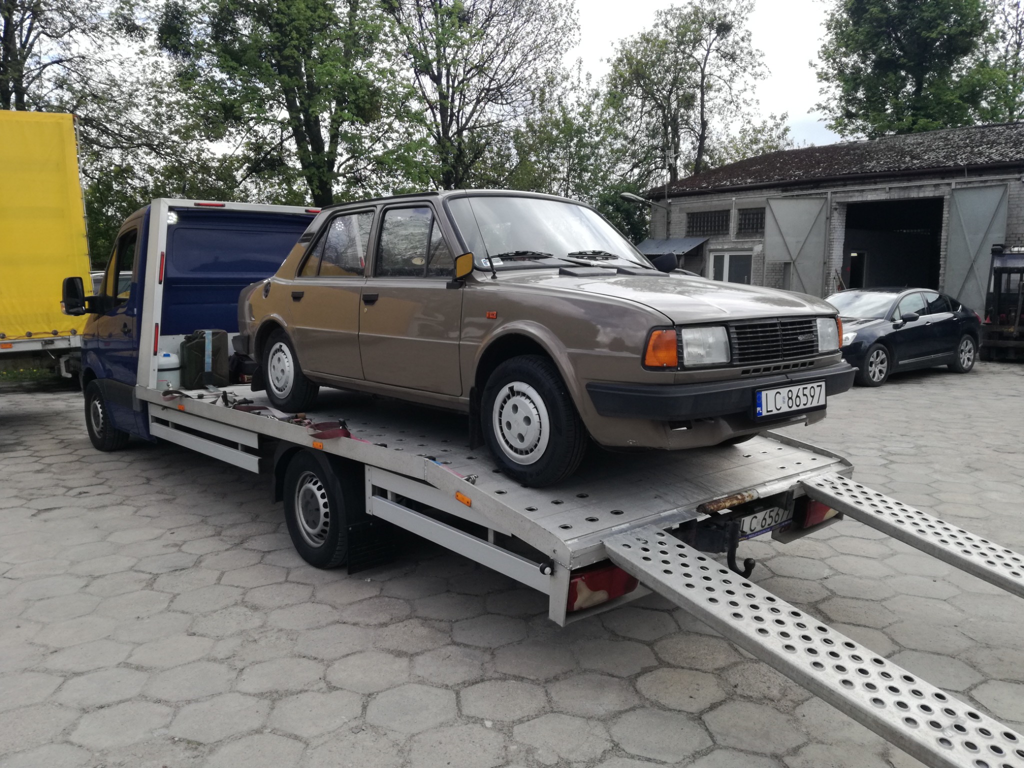 Brązowy samochód Skoda na srebrnej platformie lawety z rozłożonymi najazdami na betonowym placu, widoczny budynek z szarymi bramami w tle.