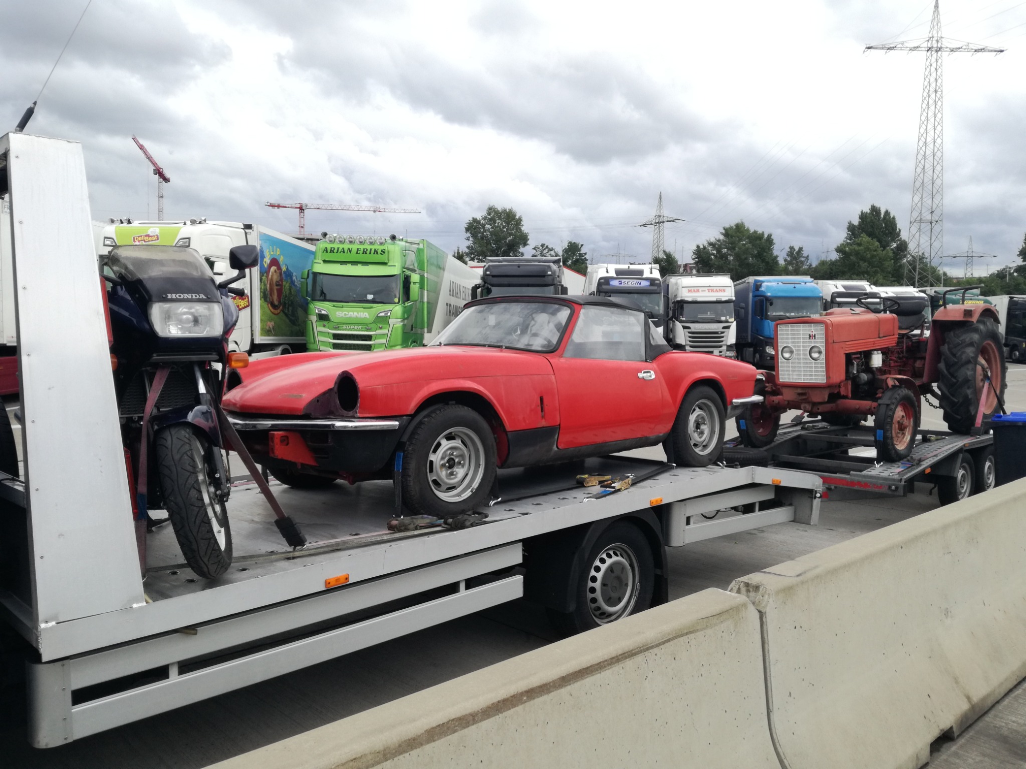 Na lawecie zaparkowany czerwony sportowy kabriolet, motocykl i stary czerwony traktor na tle zaparkowanych ciężarówek i pochmurnego nieba.