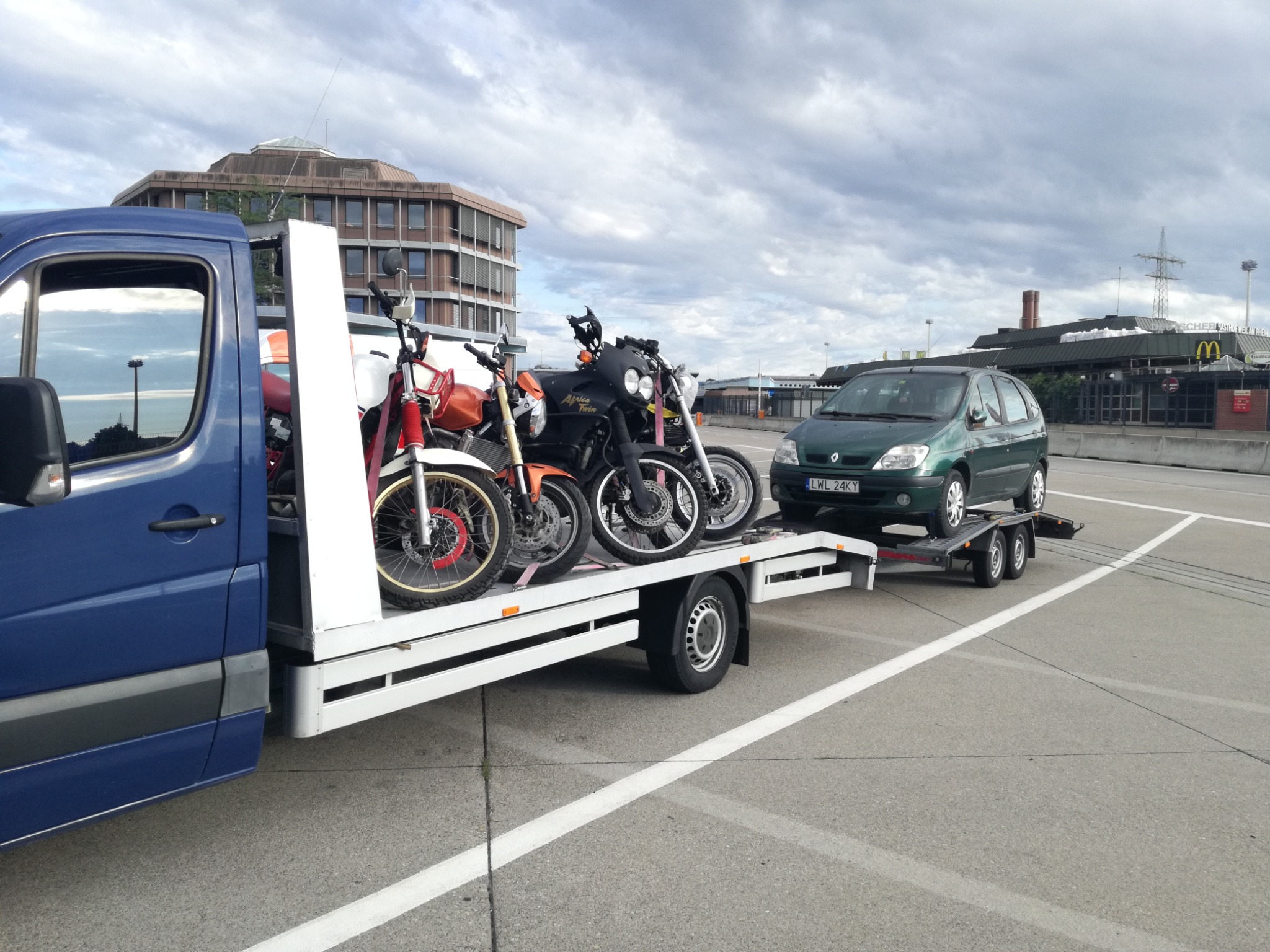 Niebieski van z platformą przewozi trzy motocykle, a za nim samochód na przyczepie, na tle budynku biurowego i pochmurnego nieba.
