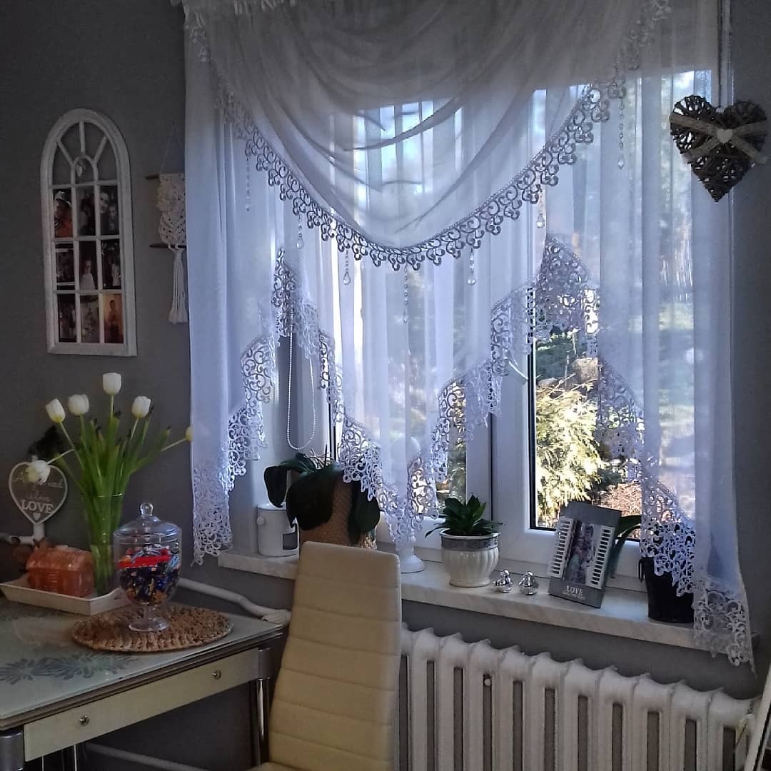 Jasne wnętrze z oknem ozdobionym delikatną, białą firanką z koronkowymi detalami i kryształkami, na parapecie doniczki z zielonymi roślinami i ramka ze zdjęciem, obok stół z wazonem tulipanów.