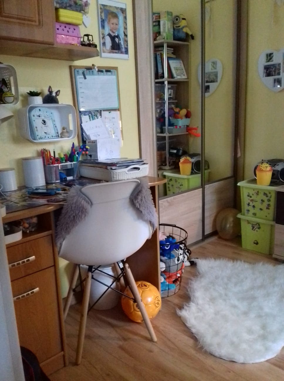 Pokój dziecięcy z biurkiem, krzesłem z futrzanymi poduszkami, regałem z zabawkami i lustrzaną szafą w tle.