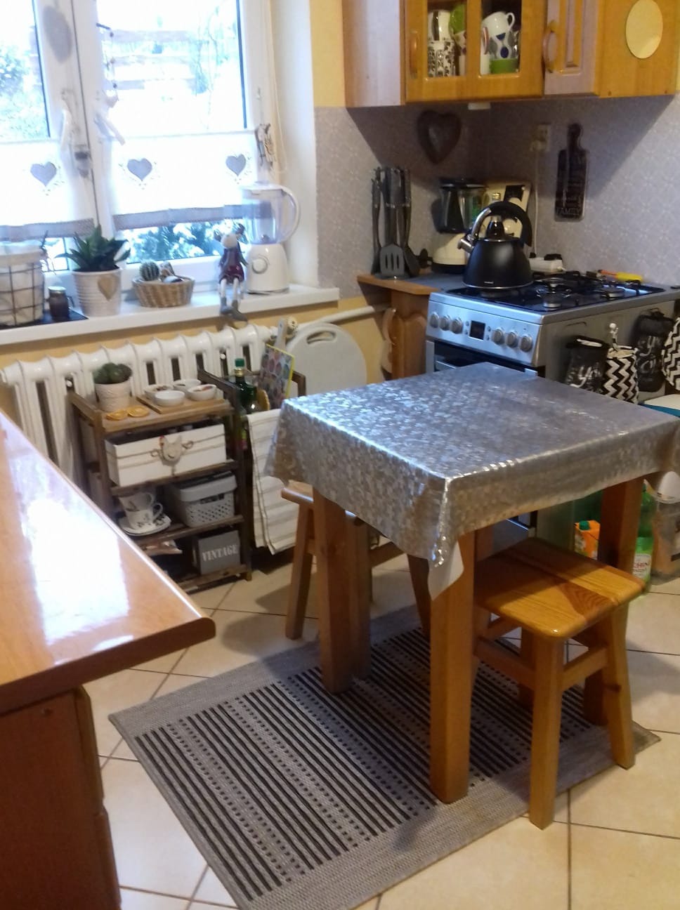 Ustawiony stół w kuchni z krzesłami, nakryty błyszczącym obrusem, widok na okno z dekoracjami w kształcie serc.