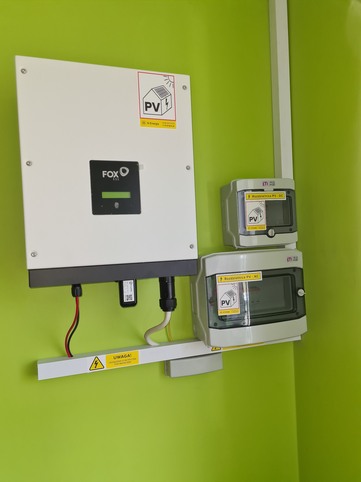 Biały inwerter FOXESS zamontowany na zielonej ścianie, obok dwie szare rozdzielnice PV-DC i PV-AC z naklejkami ostrzegawczymi, widoczny żółty znak ostrzegawczy o napięciu.