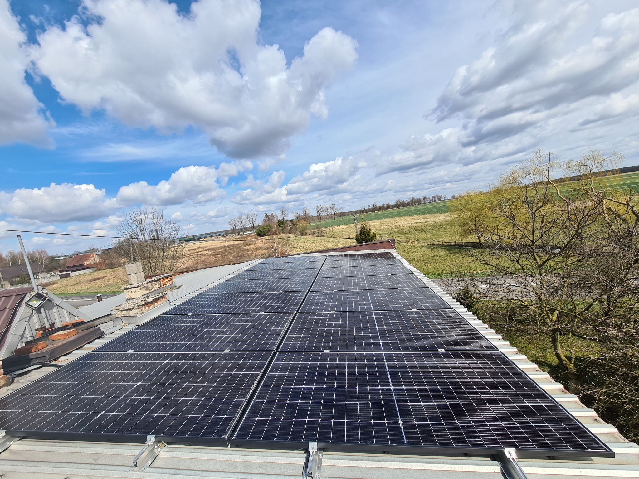 Panele słoneczne na dachu domu z widokiem na pola i chmurne niebo, uchwycone z perspektywy lekko z góry.