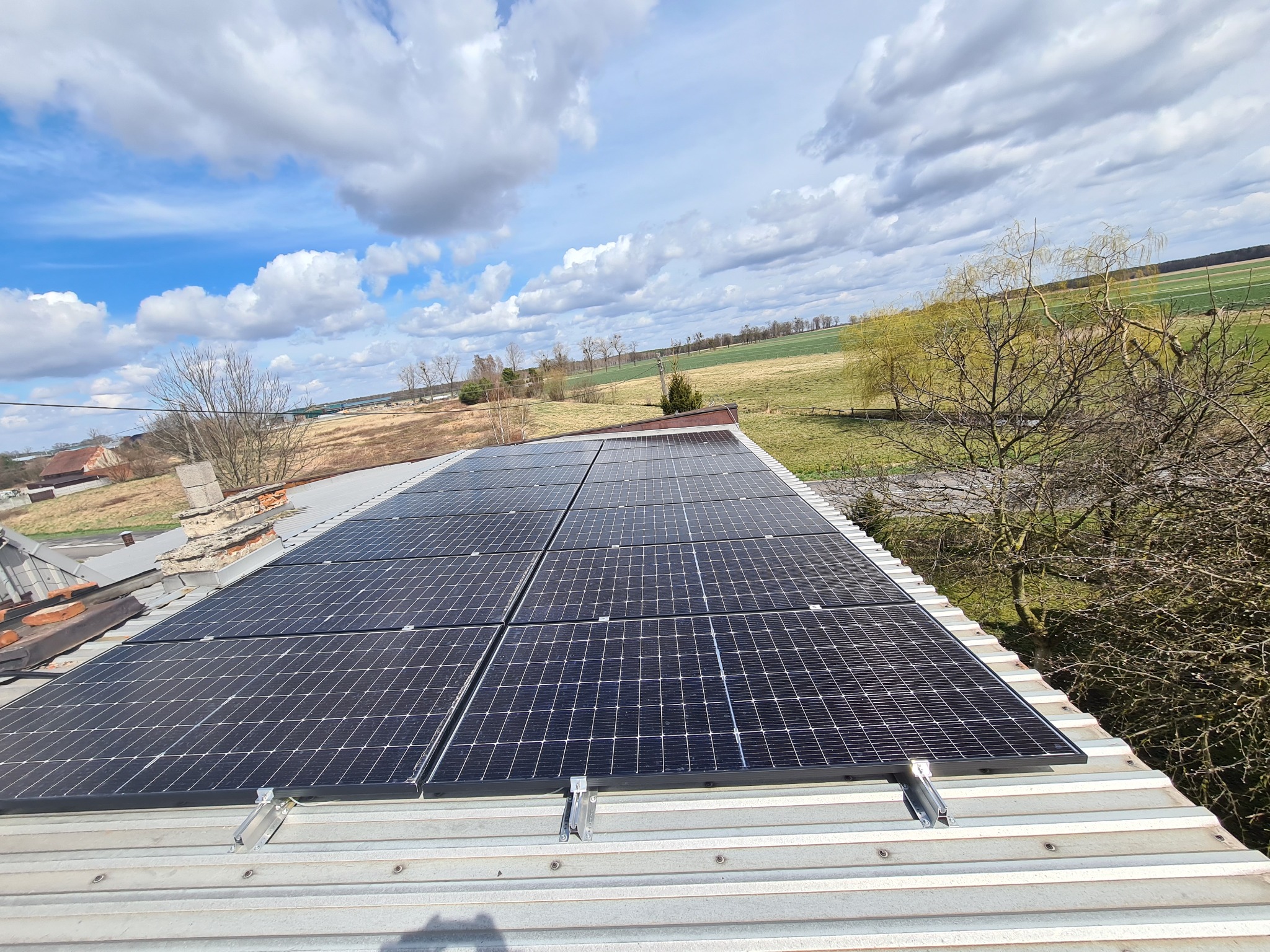 Panele słoneczne zamontowane na dachu w Oleśnicy, widok na okoliczne pola i częściowo zachmurzone niebo.