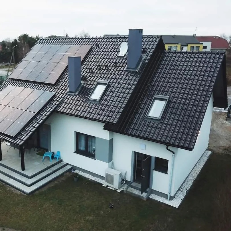 Nowoczesny dom jednorodzinny z panelami fotowoltaicznymi na dachu pokrytym ciemną dachówką, widoczne dwa kominy, okna dachowe, klimatyzator przy ścianie, mały taras z boku budynku.