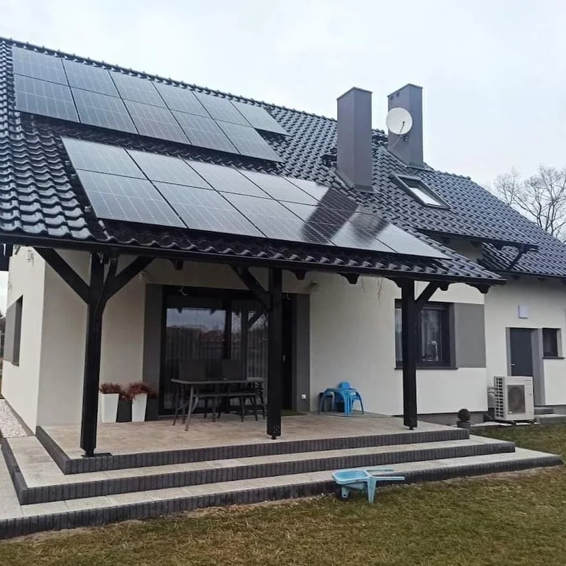 Dom jednorodzinny z panelami słonecznymi na dachu z czarnej dachówki, widoczny klimatyzator na elewacji, taras z ciemnymi filarami i schodami z kostki brukowej, błękitny wózek dziecięcy na trawniku.