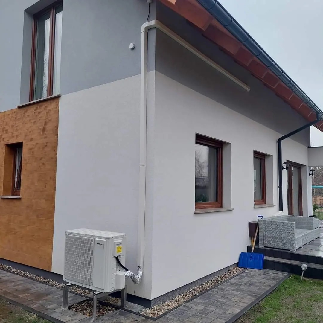 Biała pompa ciepła Viessmann zamontowana na metalowej konstrukcji przy ścianie domu z elementami ocieplenia w kolorze drewna, widoczna niebieska łopata do śniegu i szare meble ogrodowe.