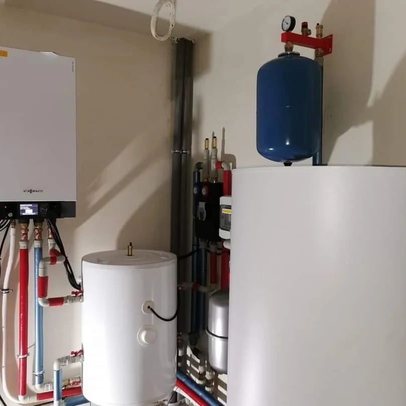 Instalacja grzewcza z białym zasobnikiem ciepłej wody, niebieskim zbiornikiem wyrównawczym i kotłem wiszącym Viessmann, widoczne liczne rury w kolorach czerwonym i niebieskim.