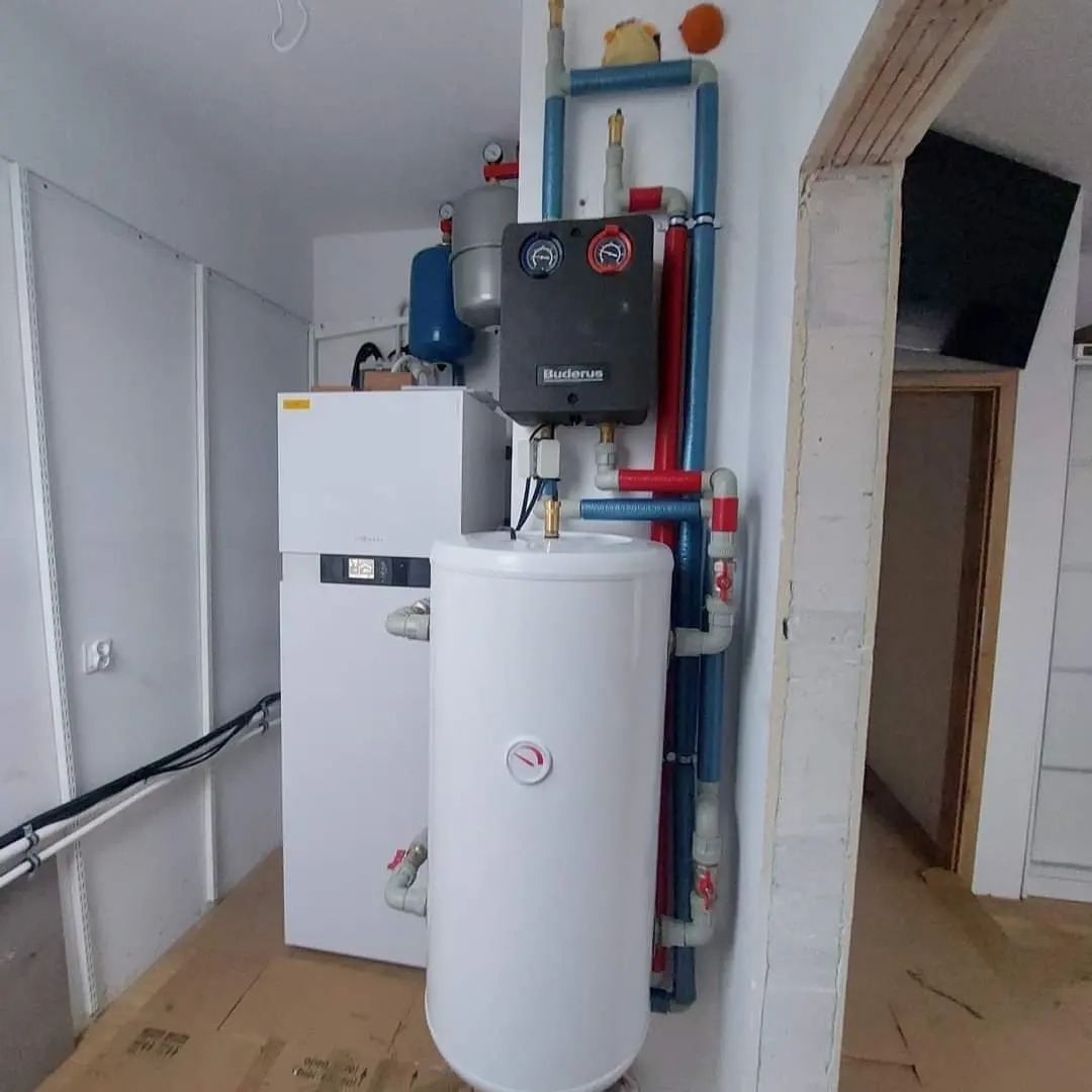 Zestaw urządzeń grzewczych Buderus: biały zasobnik ciepłej wody, szary panel sterowania, biały piec, oraz instalacja rur w kolorach niebieskim i czerwonym. Na górze rur pluszowa zabawka.