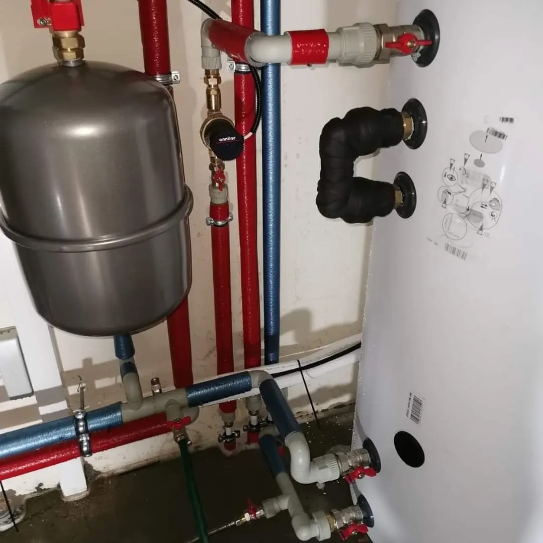 Instalacja z szarym zbiornikiem wyrównawczym, rurami w kolorze czerwonym i niebieskim oraz białym zasobnikiem ciepłej wody z czarną izolacją termiczną na przyłączach