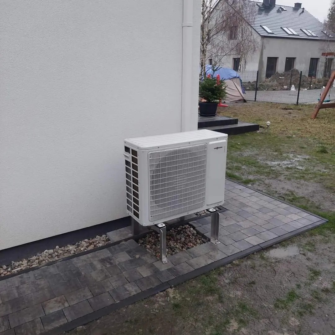 Zewnętrzna jednostka pompy ciepła marki Viessmann, zamontowana na metalowych wspornikach przy białej ścianie budynku, otoczona szarą kostką brukową i drobnymi kamieniami.