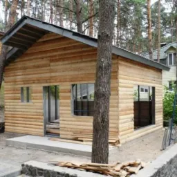 Surowy budynek z bali w trakcie budowy, z widocznymi ramami okiennymi i drzwiami, otoczony drzewami i innymi budynkami.