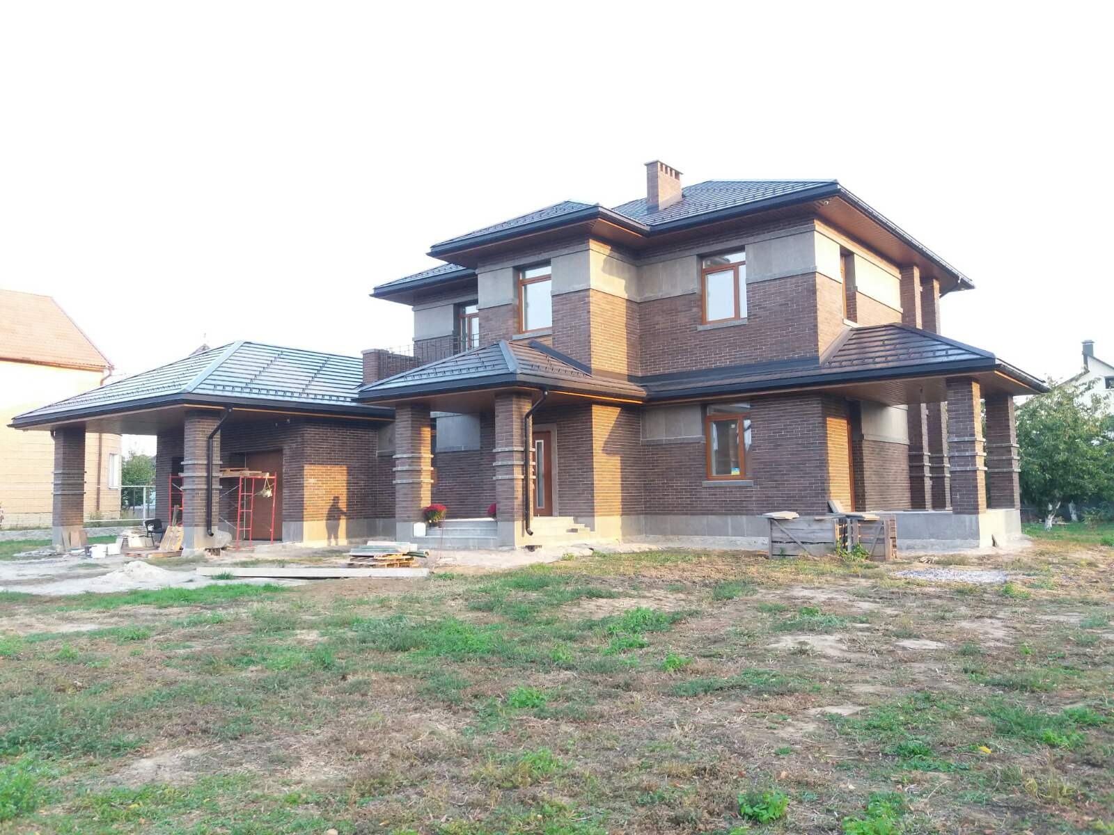 Dwukondygnacyjny dom z brązowej cegły z szarymi elementami wykończeniowymi, widoczne rusztowanie i materiały budowlane na posesji, trawnik w stanie surowym.
