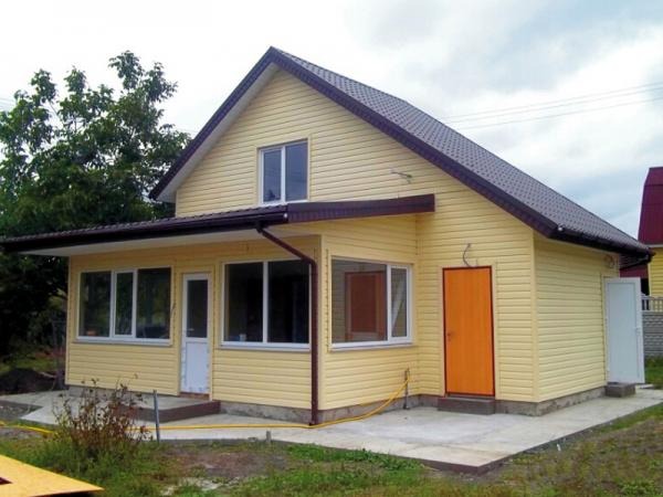 Żółty dom z brązowym dachem, wykończony sidingiem, z dobudowaną częścią i pomarańczowymi drzwiami bocznymi.